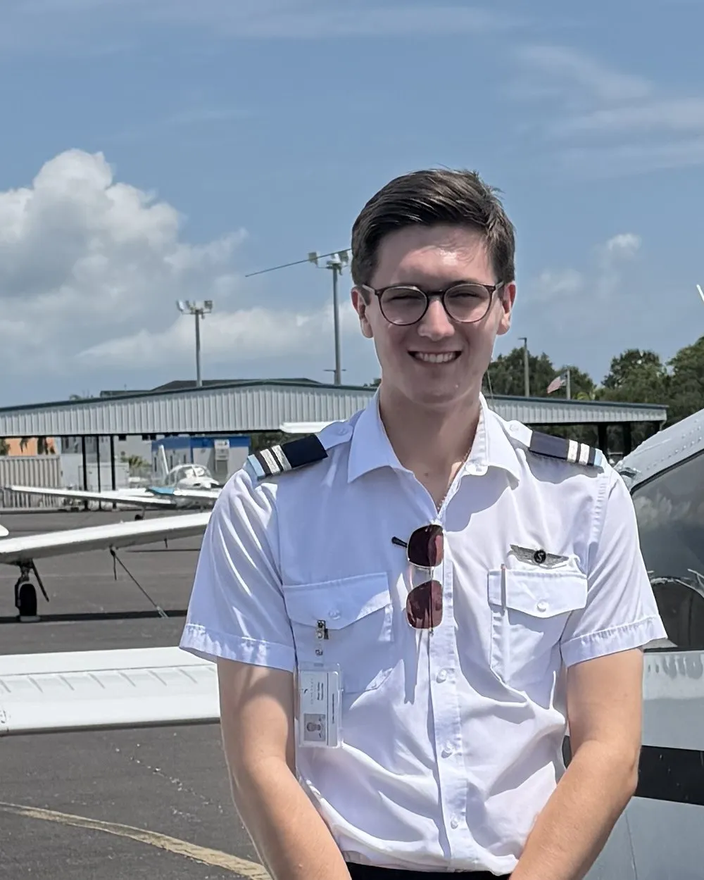 Reef smiling in a white pilot shirt with epaulettes, standing beside the wing of a training aircraft on a sunny apron